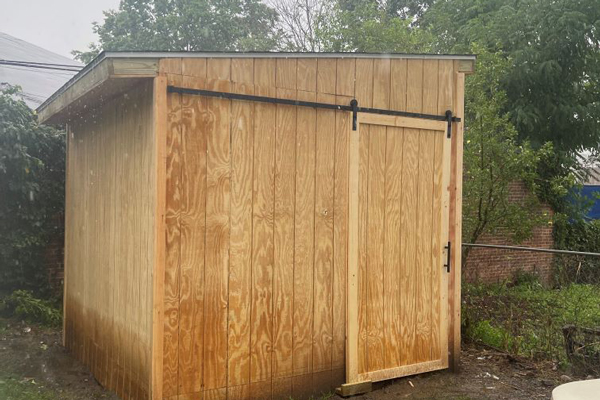 A wooden shed featuring a door and a bench positioned in front, surrounded by greenery and natural light.